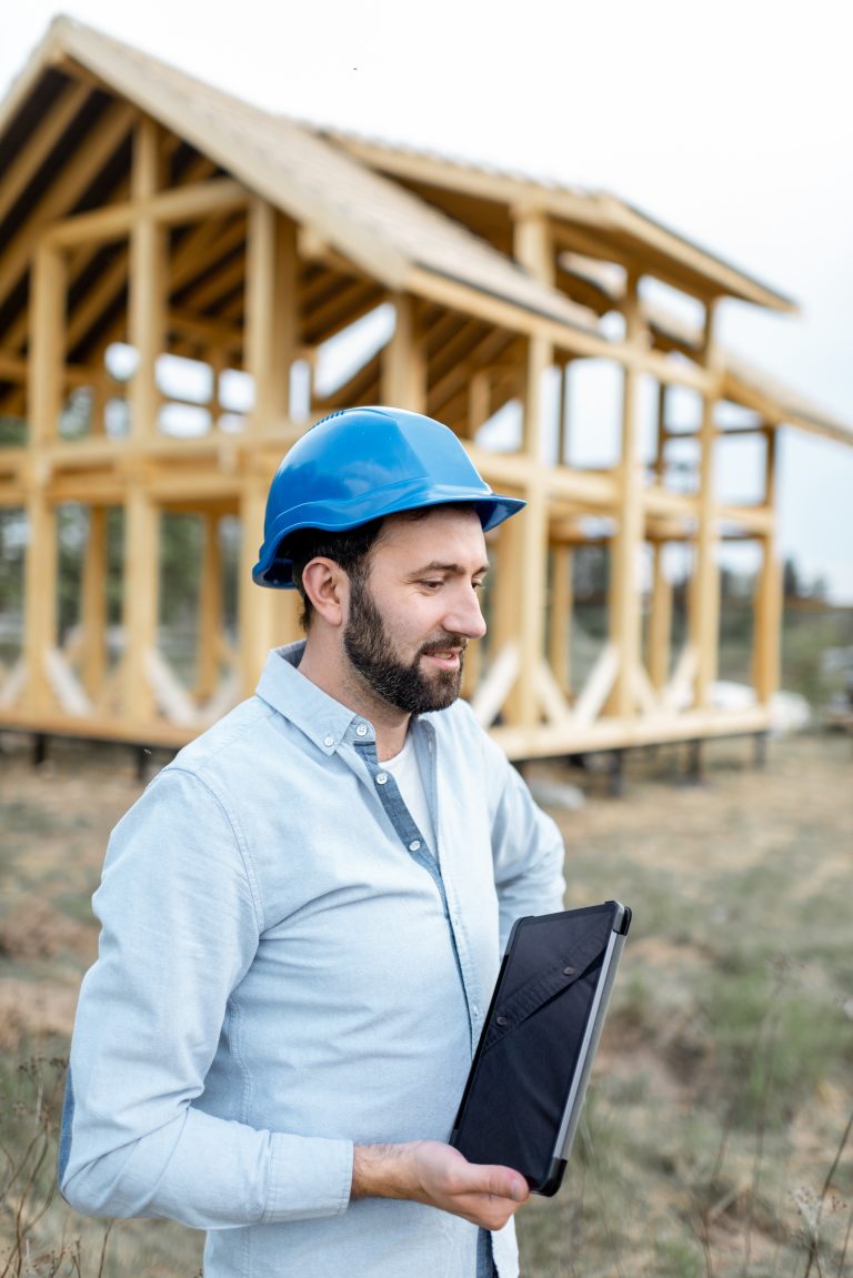 Portrait of an architect or builder with digital touchpad in front of the wooden house structure. Building and designing wooden frame house